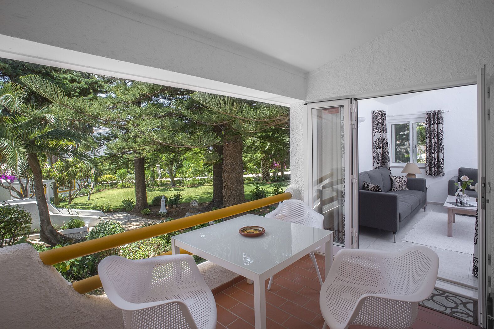 A balcony with a white table and chairs, a grey sofa, and a view of a garden with trees and a lawn.