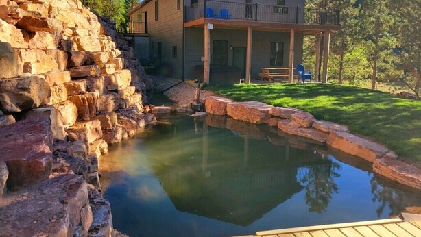 Una casa con un muro de contención de piedra, una terraza de madera y un estanque.