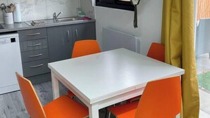 Een kleine keuken met een witte tafel en vier oranje stoelen, een grijs aanrecht en een raam met gele gordijnen.