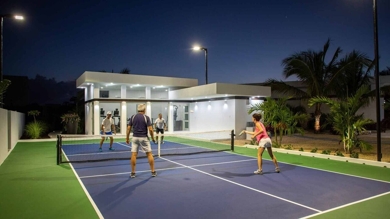 Cancha de tenis exterior con red, rodeada de palmeras y un edificio moderno con ventanas de vidrio.