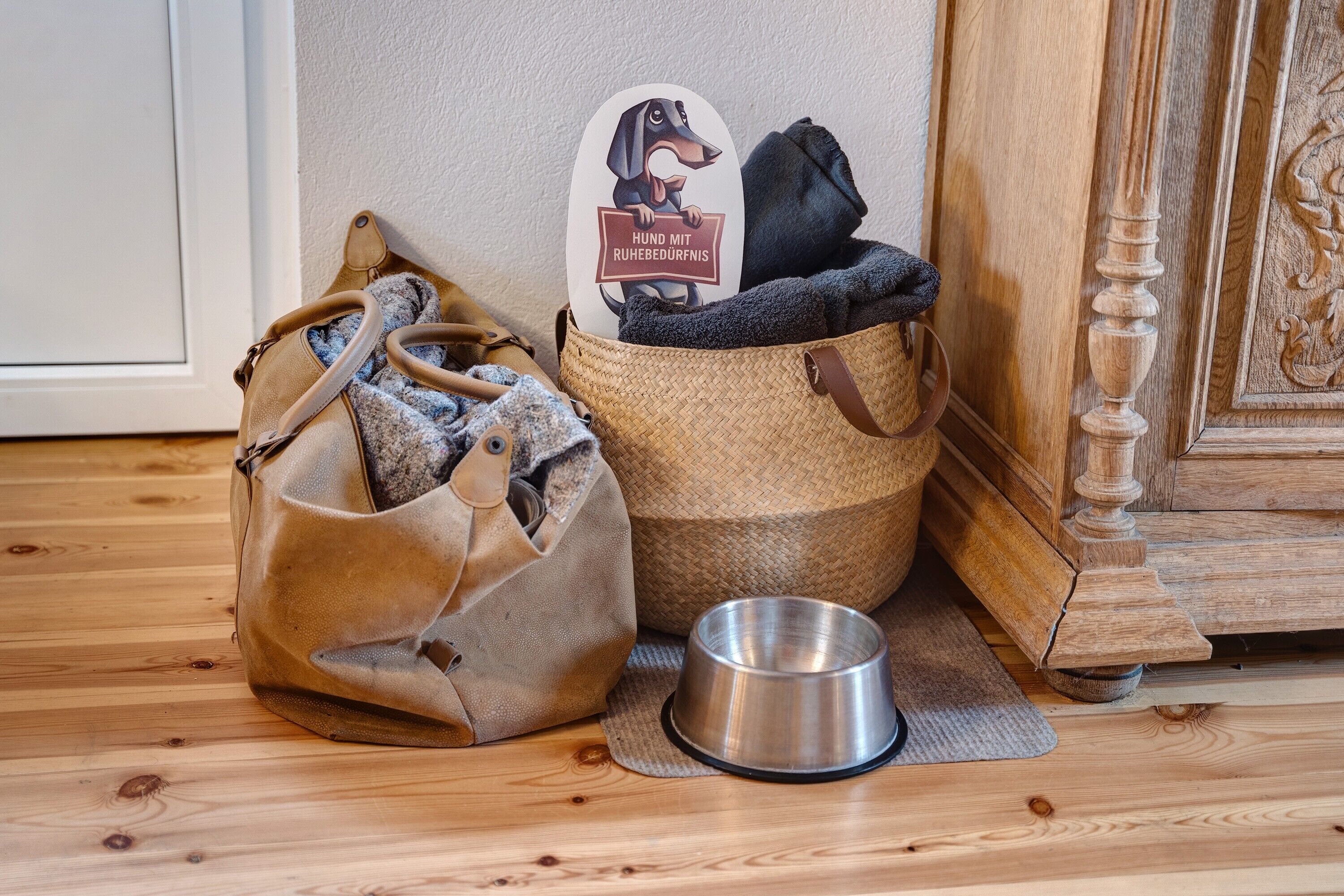 Ein Hundebett mit einem Schild, einer Metallschüssel und einer braunen gewebten Tasche auf Holzdielen.