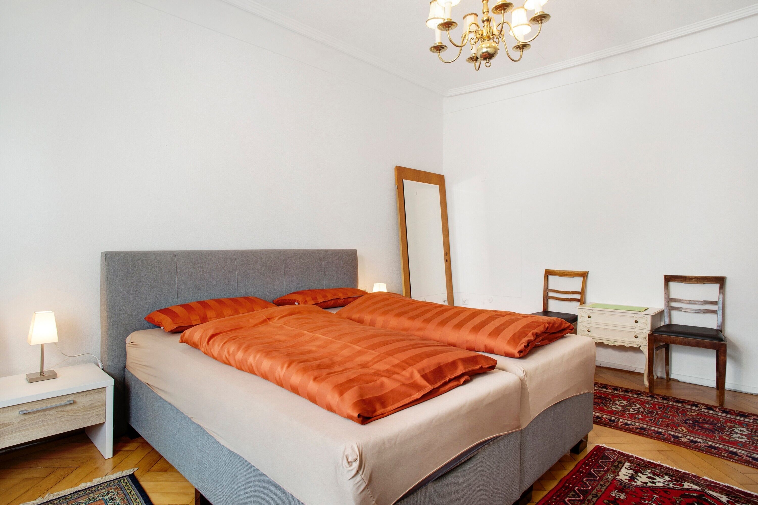 A bedroom with a bed, nightstand, dresser, mirror, two chairs, and a chandelier.