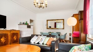 Un salon comprenant un canapé, une table ronde en bois, un téléviseur, un lustre, un miroir et une fenêtre avec des rideaux.