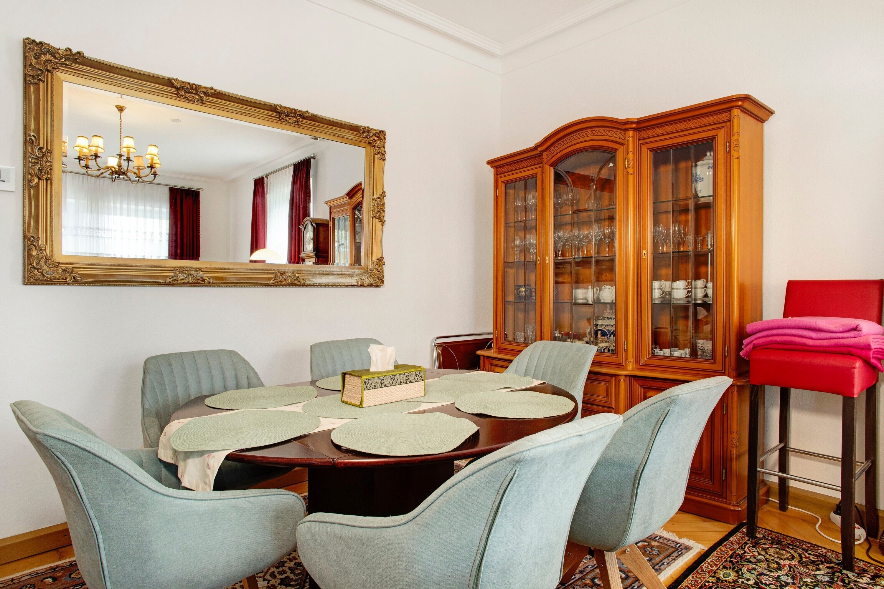 A dining room with a large mirror, a wooden cabinet with glass doors, and a red chair.
