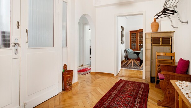 Un couloir doté d’un plancher en bois, d’un tapis rouge à motifs, d’une chaise en bois, d’une table recouverte d’une nappe en dentelle et d’un portant à vêtements suspendu.