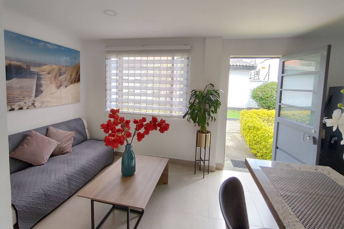 A living room with a sofa, a coffee table with a vase of red flowers, a potted plant, and a view of a garden through a window.