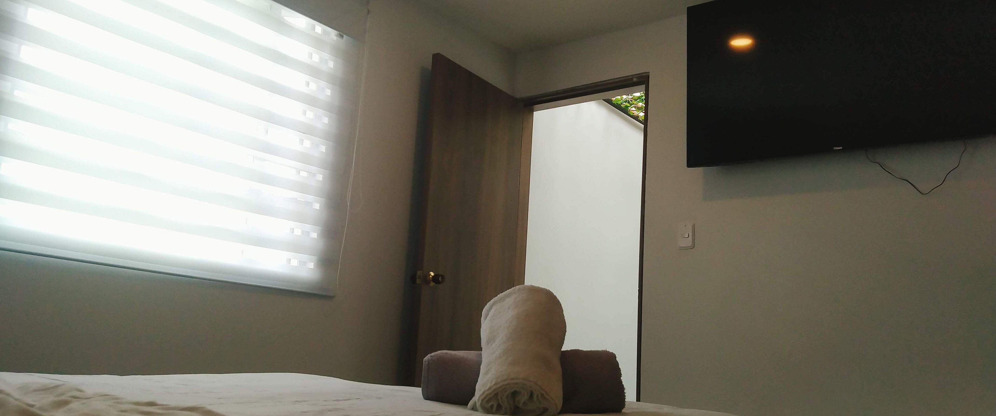 A bedroom with a bed, a window with blinds, a door, and a wall-mounted TV.