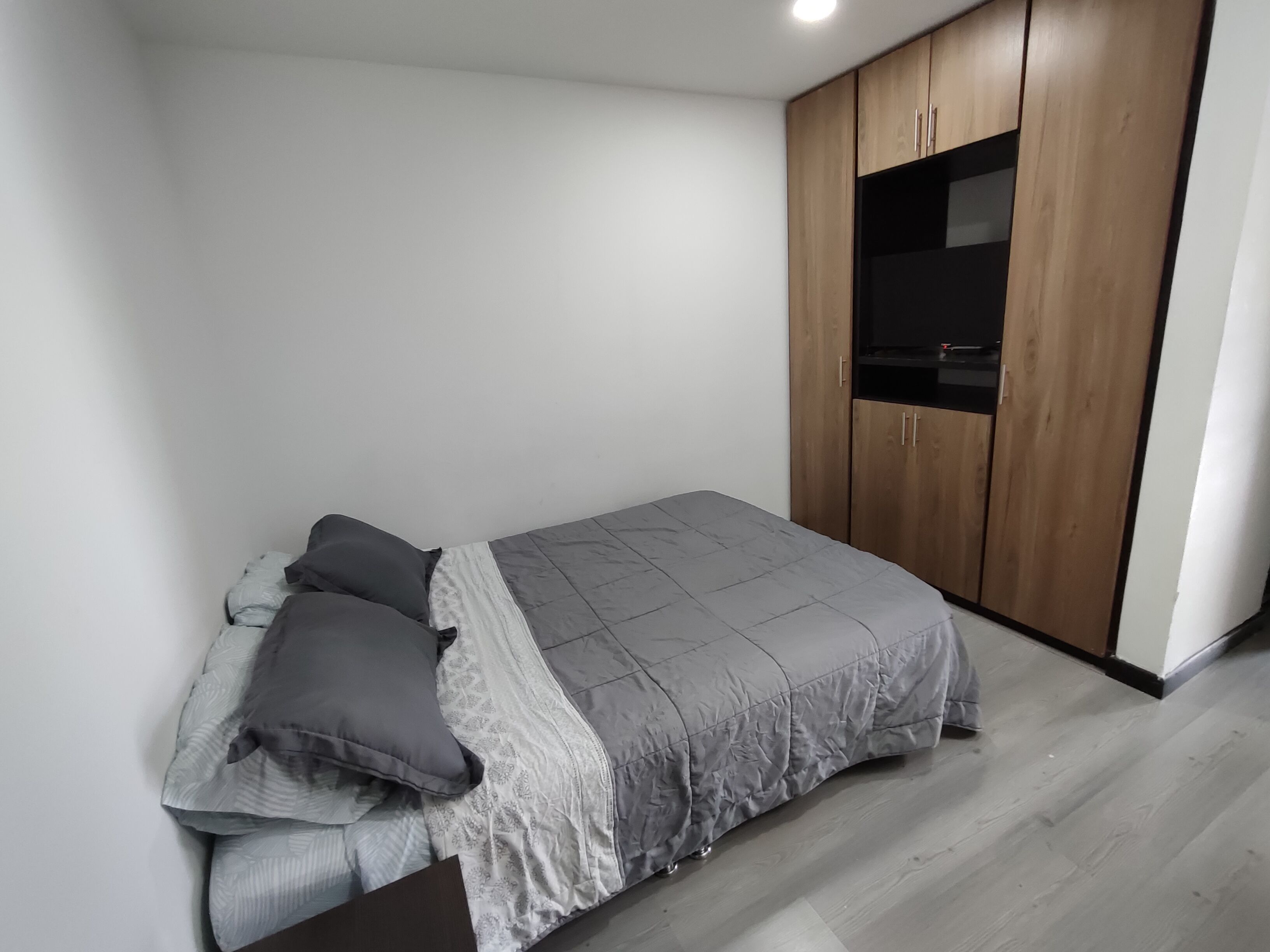 Quarto com cama, roupa de cama cinzenta, guarda-roupa de madeira e uma pequena área de cozinha.