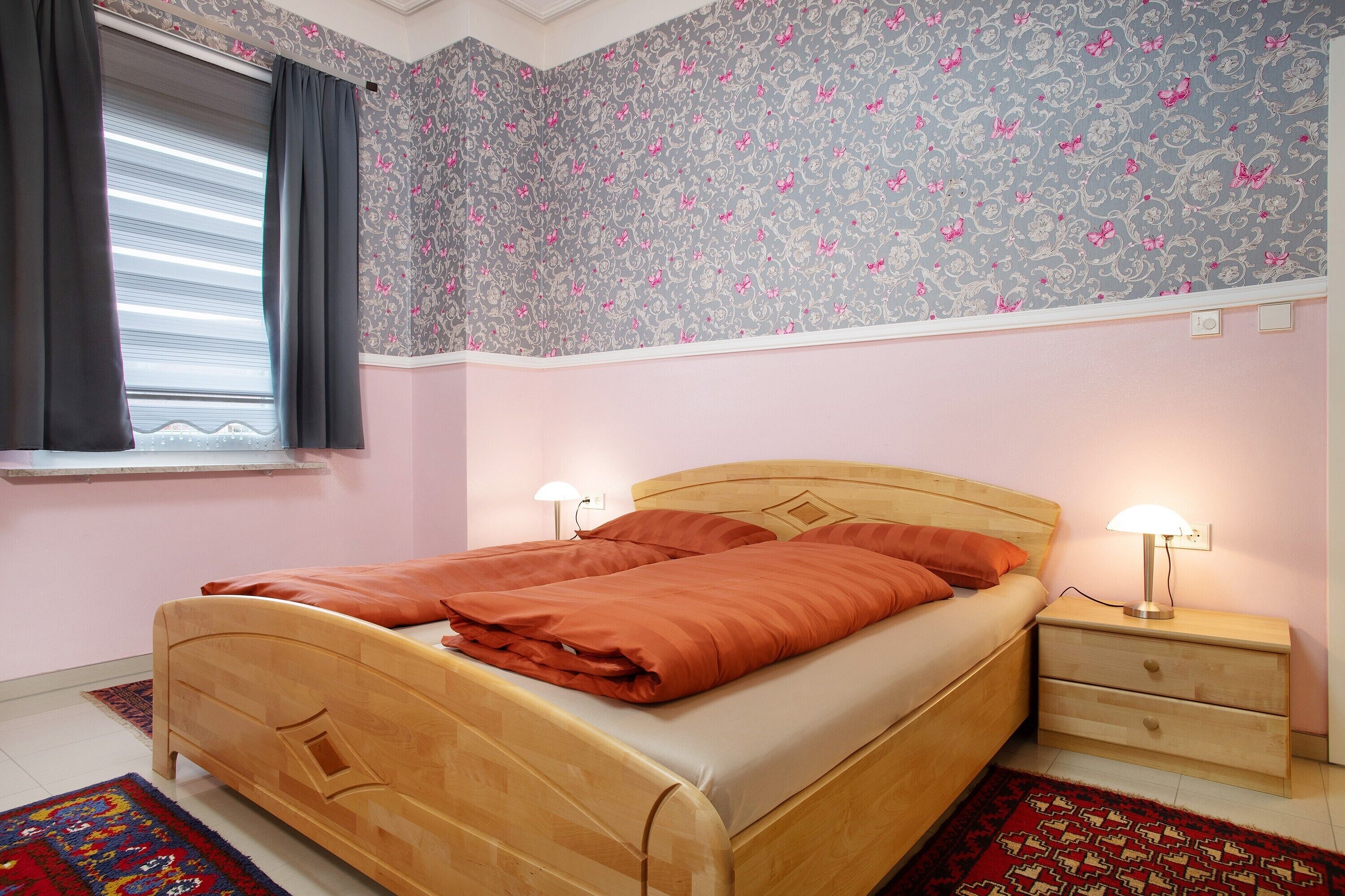 Une chambre à coucher avec un lit en bois, deux tables de chevet, une fenêtre avec des rideaux, un papier peint à motifs et un tapis rouge.
