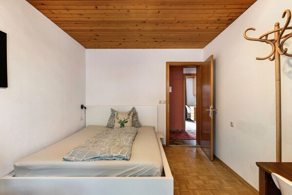 Un dormitorio con cama, techo de madera, perchero y un pasillo que conduce a otra habitación.