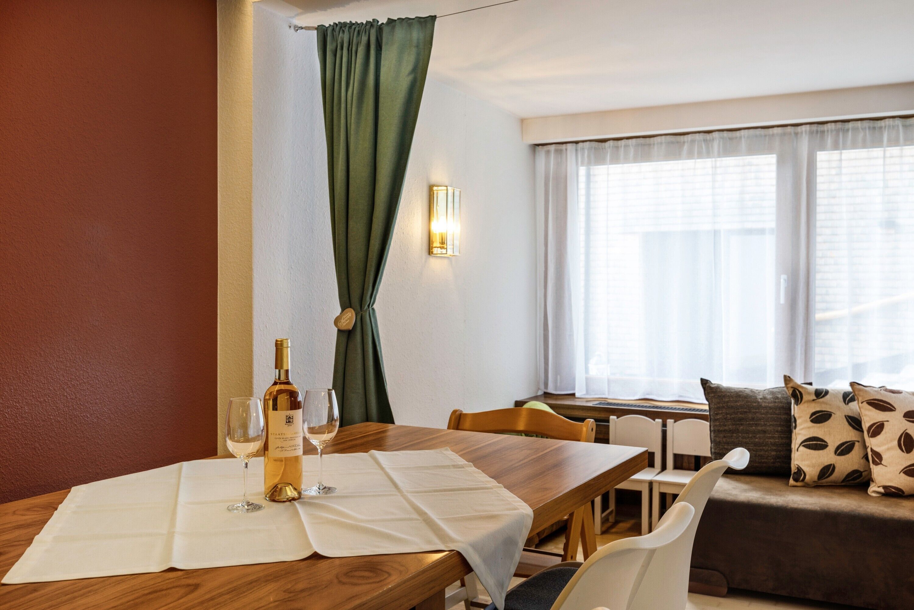Ein Essbereich mit einem Holztisch, einer Weinflasche und einem Glas auf weißer Tischdecke. Ein Fenster mit Vorhängen, eine Couch mit gemusterten Kissen und eine Wandleuchte sind ebenfalls vorhanden.