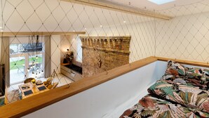 Une pièce aux hauts plafonds, dont un est en pente, avec un banc en bois, un lit recouvert d’une courtepointe à motifs floraux et une cheminée en briques.