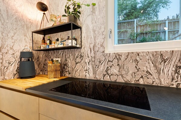 A kitchen with a black countertop, a built-in stove, a shelf with kitchen items, and a window with a view of greenery.