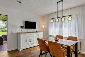 Un coin repas avec une table en bois, des chaises orange, un téléviseur posé sur un meuble blanc et des lampes suspendues.