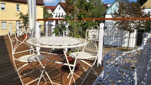 Eine Terrasse mit einem runden Tisch und weißen Metallstühlen, umgeben von einer Holzterrasse und einem Metallzaun.