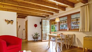 Una habitació amb sostre de fusta, una taula de dinar amb cadires, una cadira vermella, una finestra amb cortines i una porta.