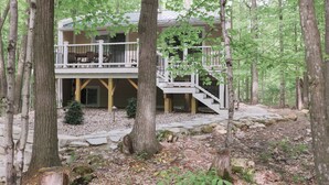 A cabin with a deck and stairs, surrounded by trees and a stone pathway. - The best patio and firepit at The Cat's Lair in the woods!  Level 2 EV charger! (Egg Harbor)