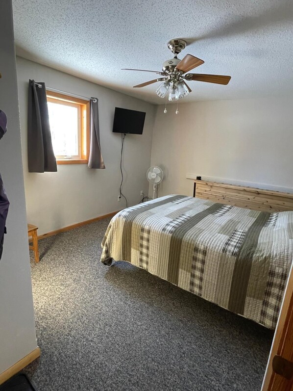 Un dormitorio con cama, ventilador de techo, televisor montado en la pared y ventana con cortinas.