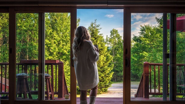 A person standing at a wooden door with glass panels, looking out at a deck with a view of a forested area. - Hot Tub, Sauna, Game Room, Fire Pit, BBQ, Karaoke, Dogs OK (Bethel)