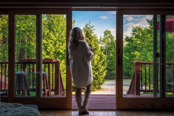 Eine Person steht vor einer Holztür mit Glasscheiben und blickt auf eine Terrasse mit Blick auf einen bewaldeten Bereich.