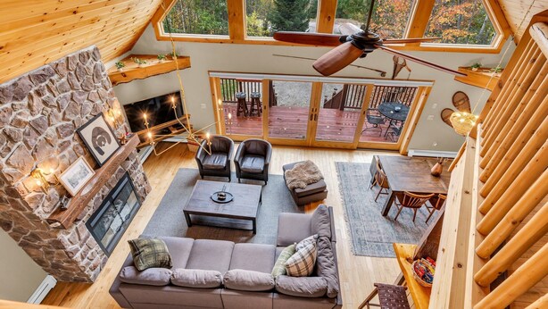 A cozy interior with a stone fireplace, a seating area with sofas and chairs, a dining table, and a view of the outdoors through large windows. - Hot Tub, Sauna, Game Room, Fire Pit, BBQ, Karaoke, Dogs OK (Bethel)