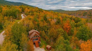 A cabin with a wooden exterior, surrounded by autumn-colored trees, with a dirt path leading to it. - Hot Tub, Sauna, Game Room, Fire Pit, BBQ, Karaoke, Dogs OK (Bethel)