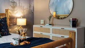 A cozy bedroom with a bed, a coffee maker, a bedside table with books, and a wall-mounted mirror reflecting a forest scene. - Hot Tub, Sauna, Game Room, Fire Pit, BBQ, Karaoke, Dogs OK (Bethel)
