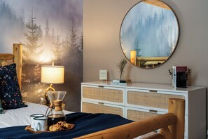 A cozy bedroom with a bed, a coffee maker, a bedside table with books, and a wall-mounted mirror reflecting a forest scene.