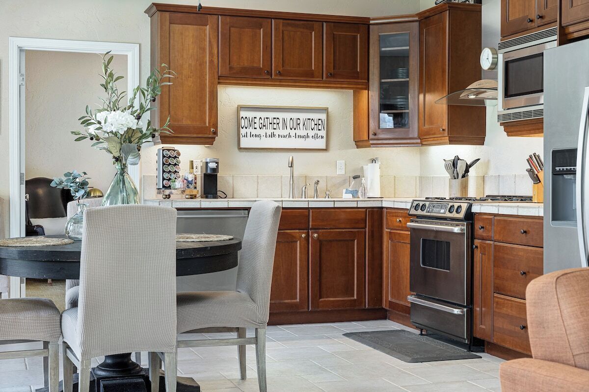 A kitchen with wooden cabinets, a dining table with chairs, a coffee station, and a clock on the wall.