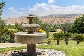 Ein steinerner Brunnen mit abgestufter Gestaltung, umgeben von Kies, Palmen und einem Bergpanorama.