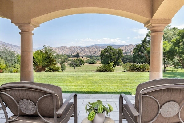 A covered outdoor seating area with two chairs and a potted plant, overlooking a well-maintained lawn and distant mountains.