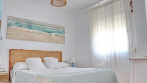 A bedroom with a bed, a bedside table, a lamp, a window with curtains, and a beach-themed painting on the wall. - Apartment Virtudes III (Conil de la Frontera)