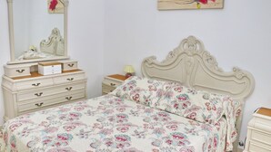 Un dormitorio con cama, tocador, mesita de noche, espejo y una obra de arte floral en la pared.