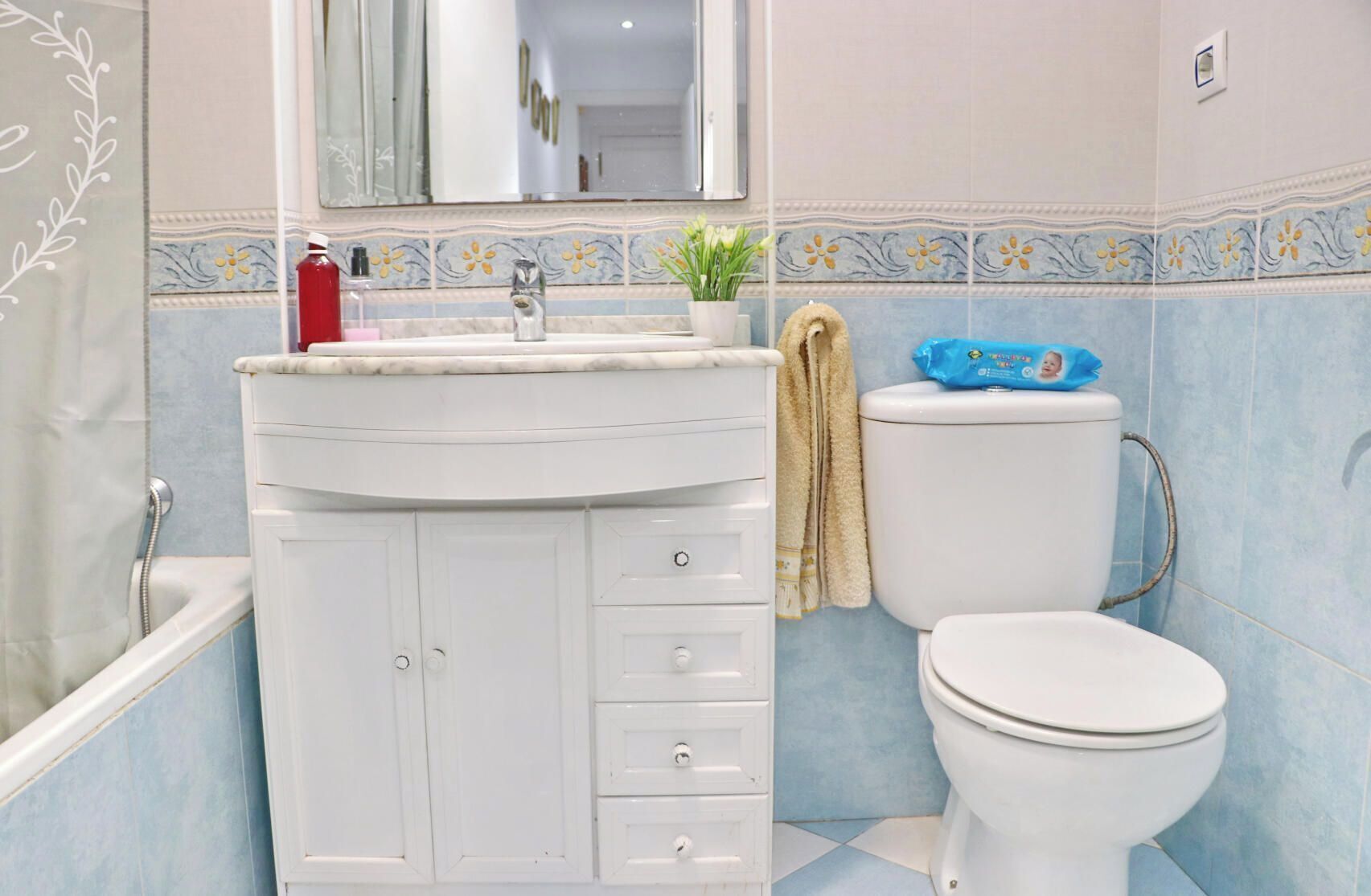 A bathroom with a white sink, mirror, and toilet. There is a towel hanging on the wall and a small potted plant on the sink.