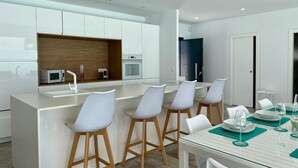 A modern kitchen with white cabinets, a central island with bar stools, and a dining area with a white table and chairs. - El Azul House in Chiclana (Chiclana)