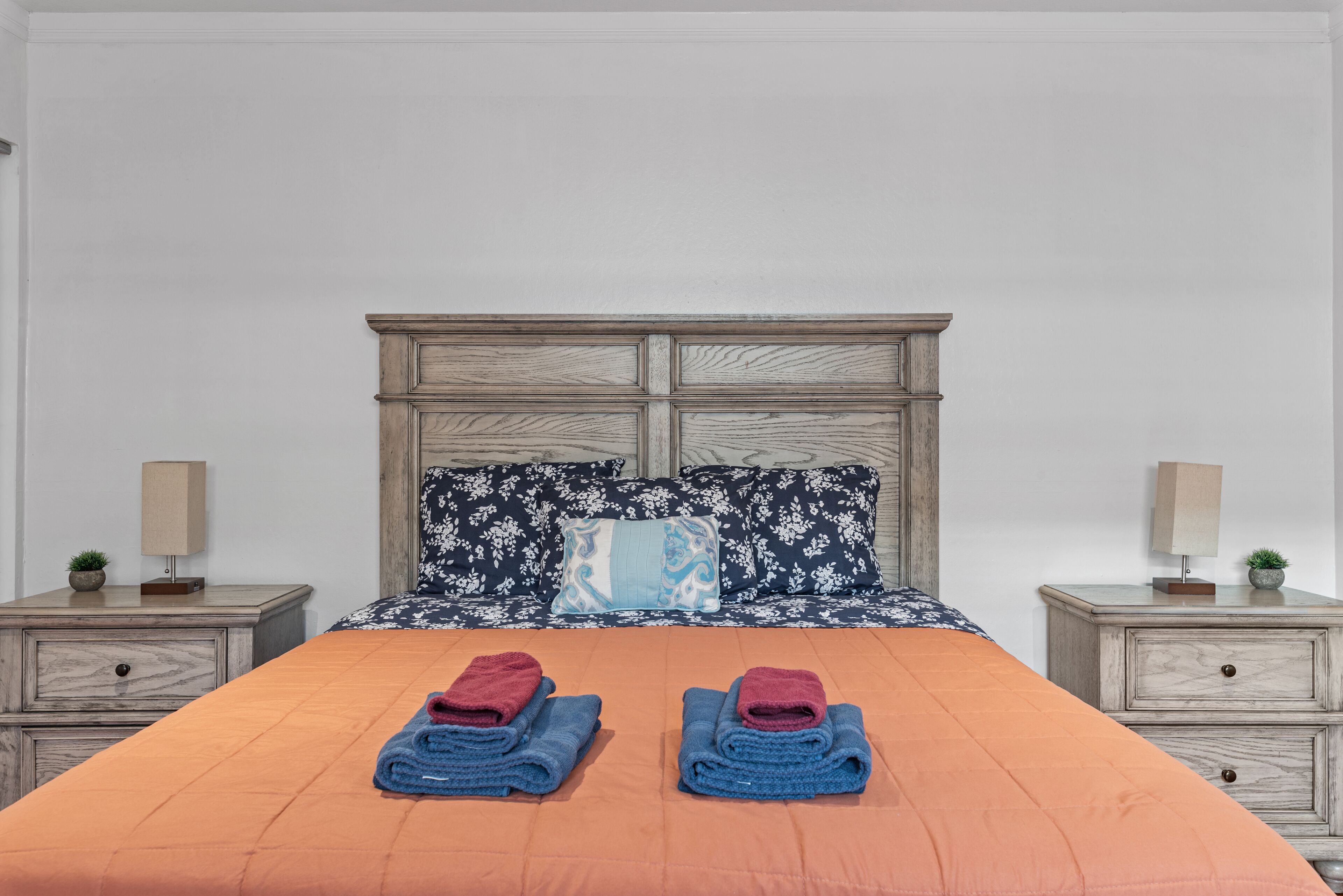 Un lit bien fait, avec deux serviettes pliées, une tête de lit en bois et deux tables de chevet assorties, sur lesquelles sont posées des lampes et de petites plantes.