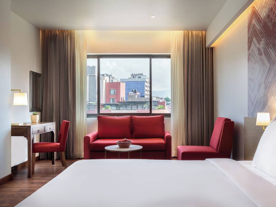 Una habitación de hotel con una cama grande, un sofá rojo, un escritorio con lámpara y vistas a edificios a través de la ventana.