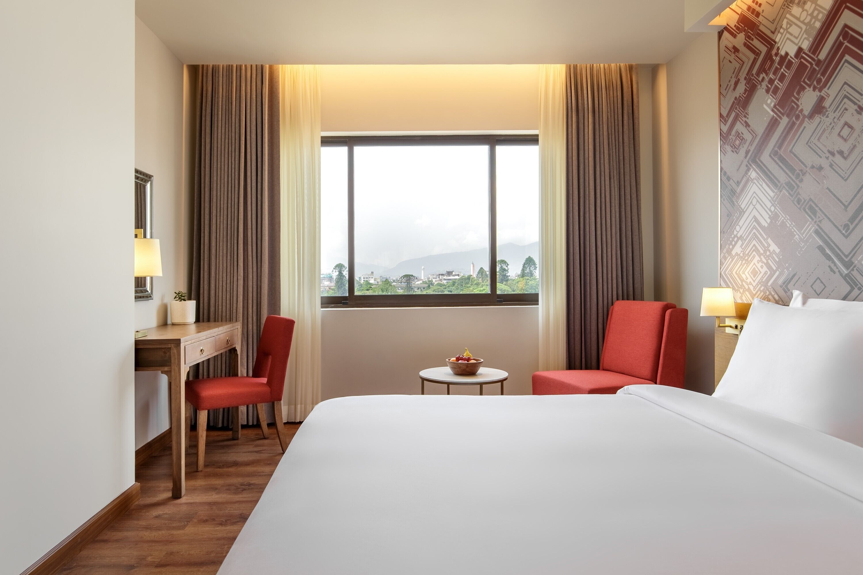 Quarto de hotel com uma cama grande, uma escrivaninha com cadeira, uma mesinha com uma tigela de frutas e vista para a cidade através de uma janela ampla.