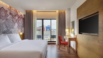 A hotel room with a large bed, a desk with a chair, a TV, and a view of the city through the window.
