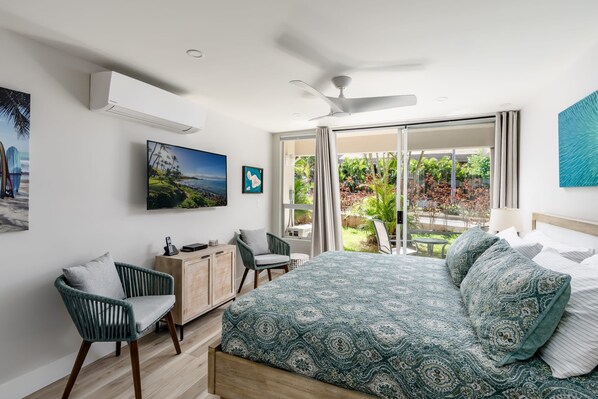 A bedroom with a bed, a desk, a chair, a ceiling fan, and a view of a garden through a sliding glass door.