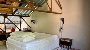 A bedroom with a white bed, a colorful rug, a small black nightstand, and a metal-framed ceiling.