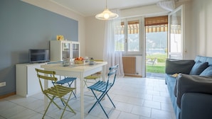 Een moderne woonkamer met een bank, een eettafel met stoelen, een televisie en een keukenhoek met kasten en een schaal fruit.