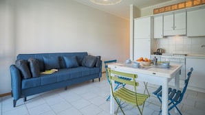 Een knus woongedeelte met een blauwe bank, een eettafel met stoelen en een kitchenette met witte kastjes.