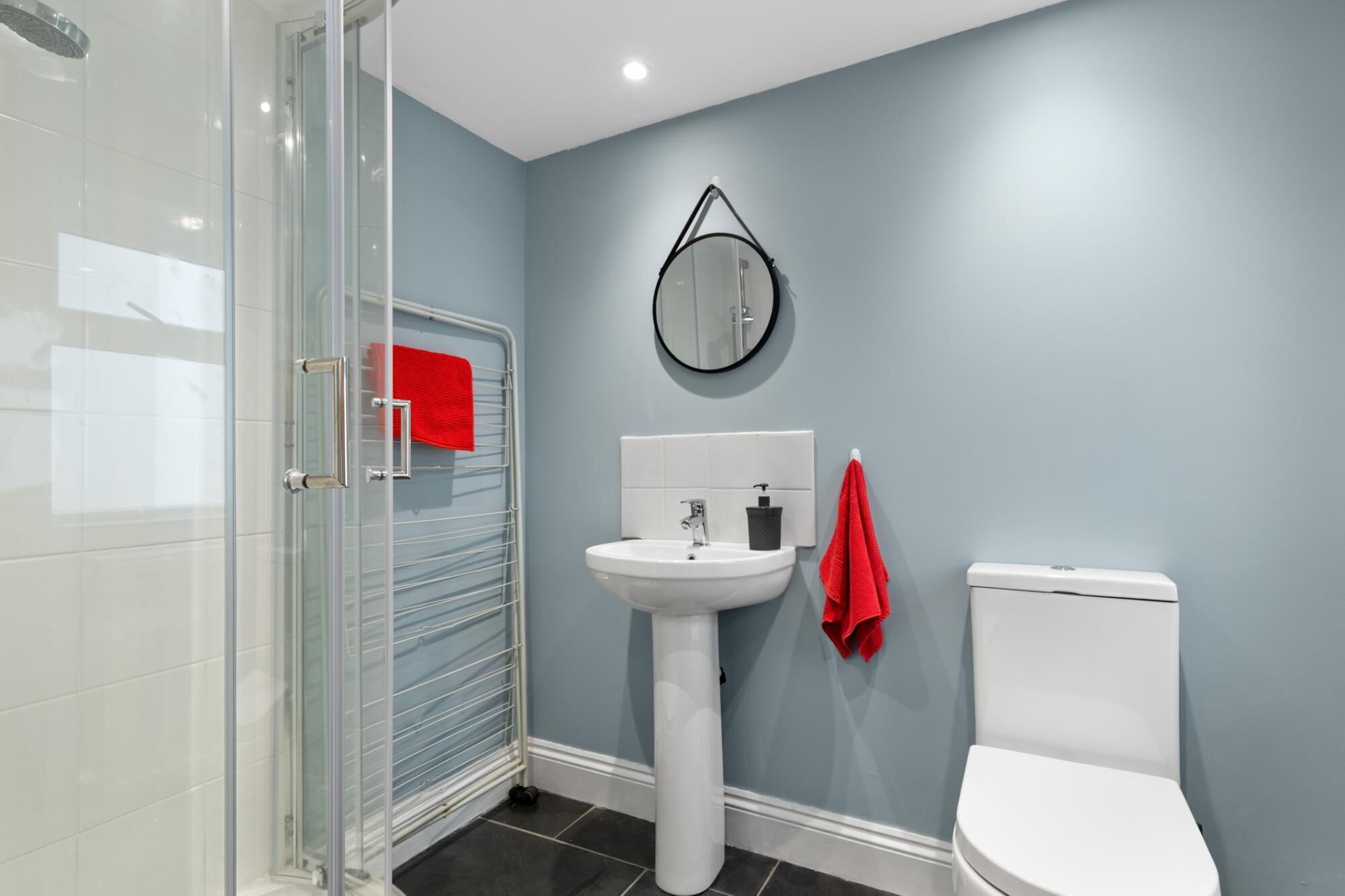 A bathroom with a glass shower enclosure, a white pedestal sink, a round mirror, and a white toilet.