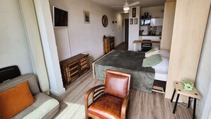 A compact living space with a bed, a sofa, a wooden chair, a small table, and a kitchenette area. - Renovated studio, sea view, close to beaches and shops... (ST MANDRIER SUR MER)
