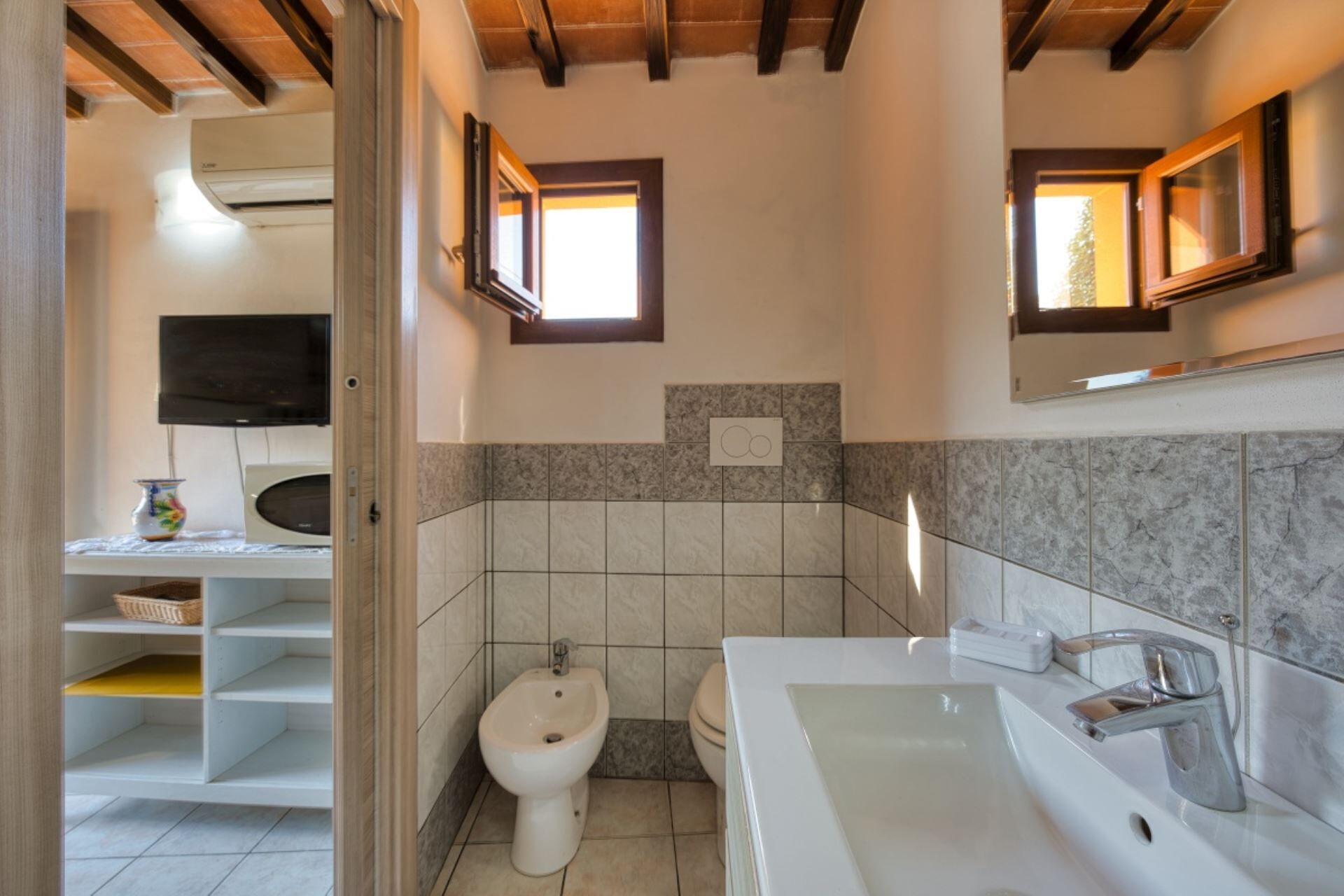 Une salle de bain comprenant des toilettes, un bidet et un lavabo avec un miroir au-dessus. On y trouve une étagère avec un panier et un téléviseur fixé au mur.
