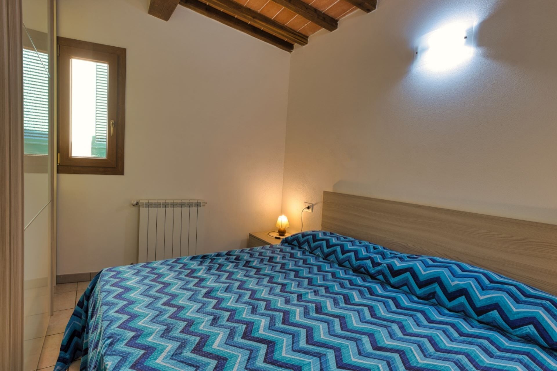 Un dormitorio con cama, mesita de noche, ventana y radiador.