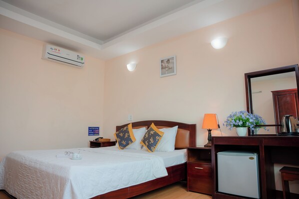 A neatly made bed with a white bedspread, two decorative pillows, a bedside table with a lamp, a small refrigerator, and a wall-mounted air conditioner.