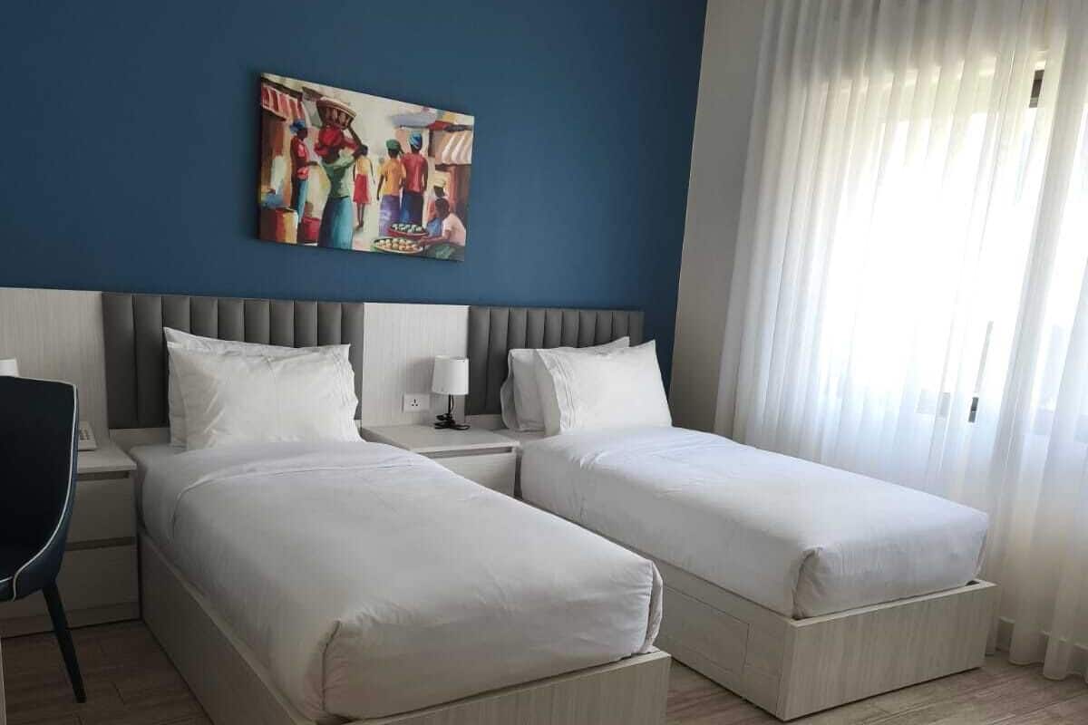 A hotel room with two single beds, a nightstand, a chair, a painting on the wall, and a large window with curtains.