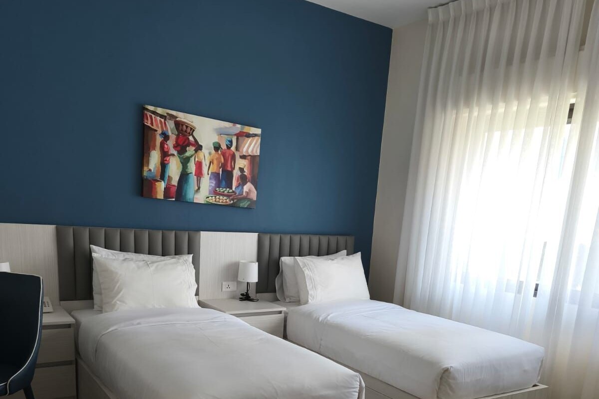 Une chambre d’hôtel avec deux lits simples, une table de chevet, une chaise, un tableau au mur et une grande fenêtre avec des rideaux.
